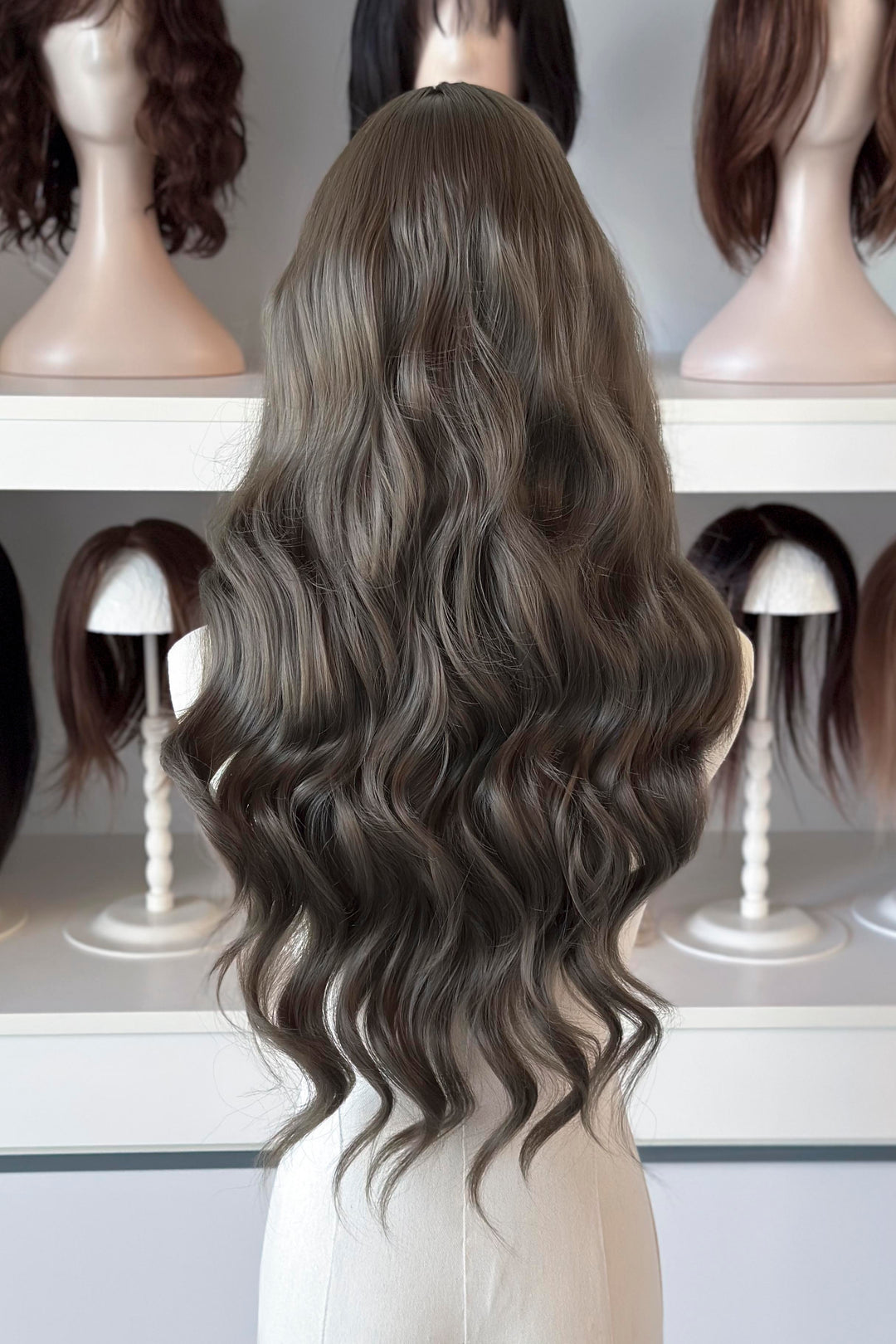 long wavy dark ash golden brown with green undertones wig in front of other mannequin heads with wigs on a shelf.