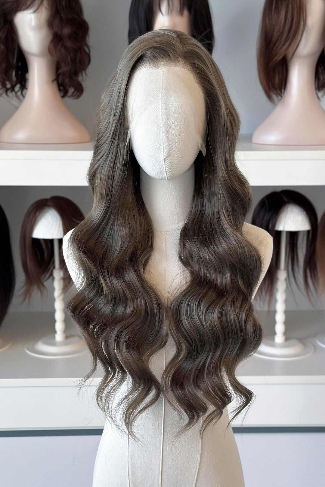 long wavy dark ash golden brown with green undertones wig in front of other mannequin heads with wigs on a shelf.