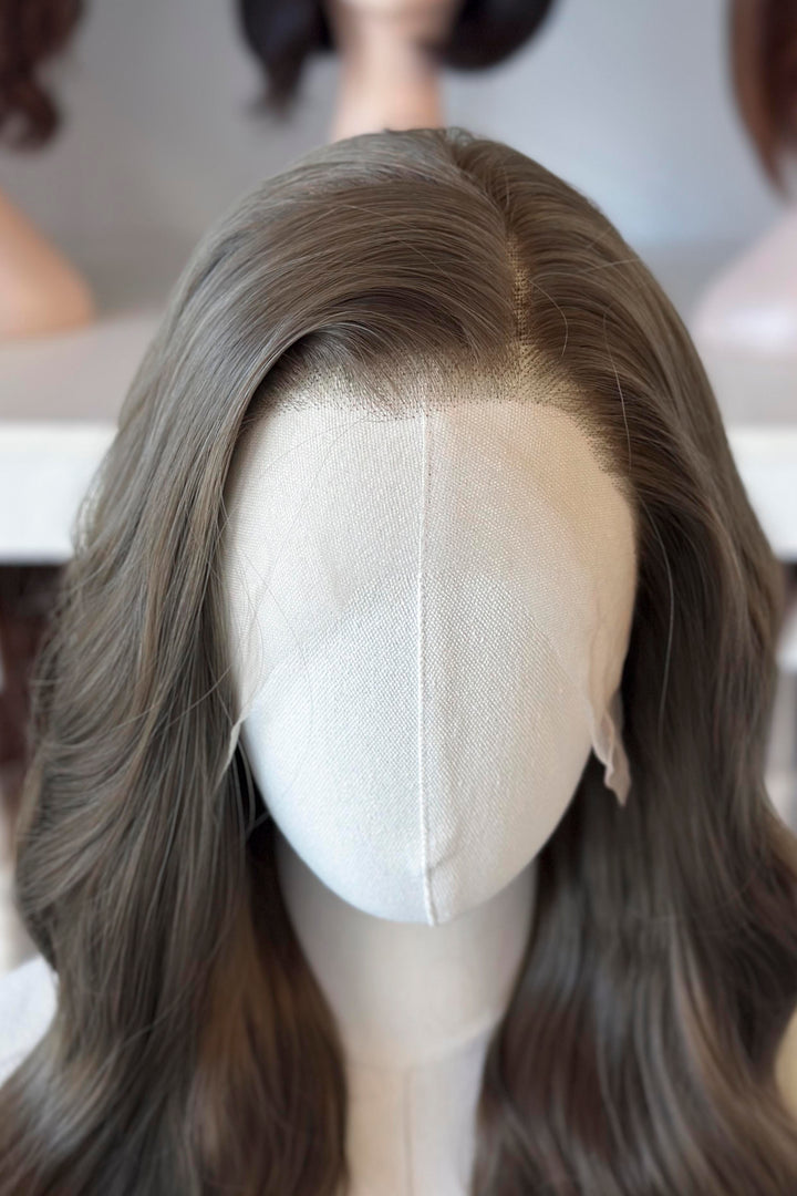 long wavy dark ash golden brown with green undertones wig in front of other mannequin heads with wigs on a shelf.