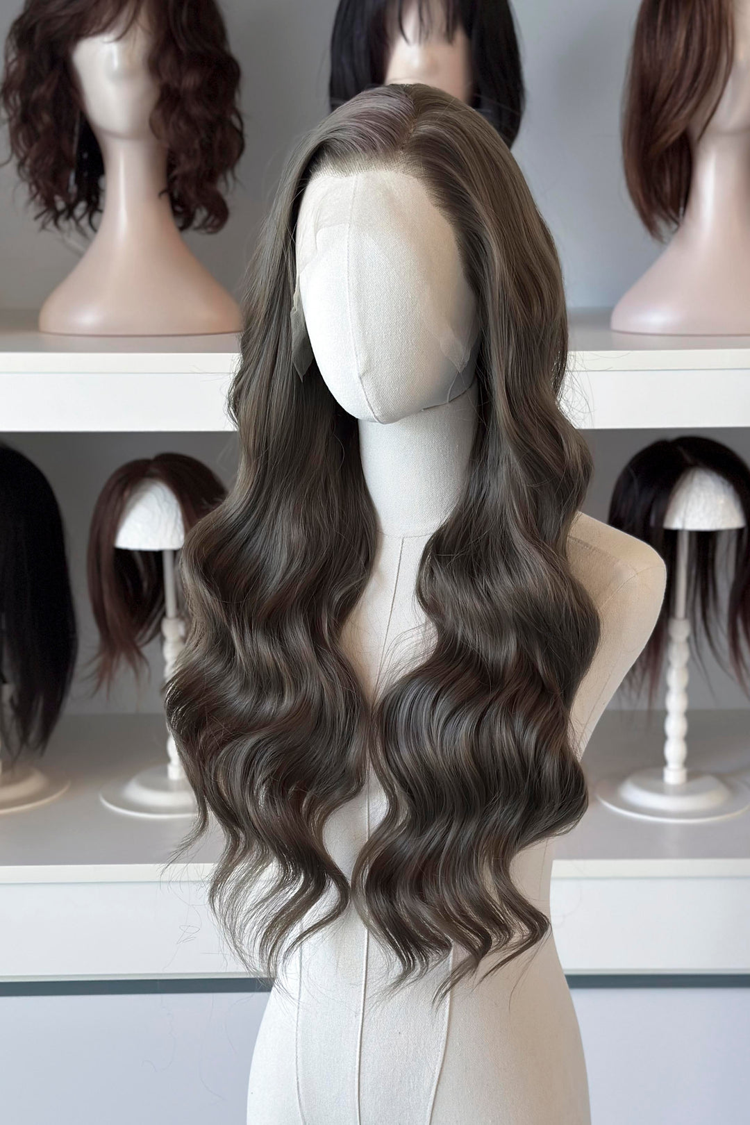 long wavy dark ash golden brown with green undertones wig in front of other mannequin heads with wigs on a shelf.