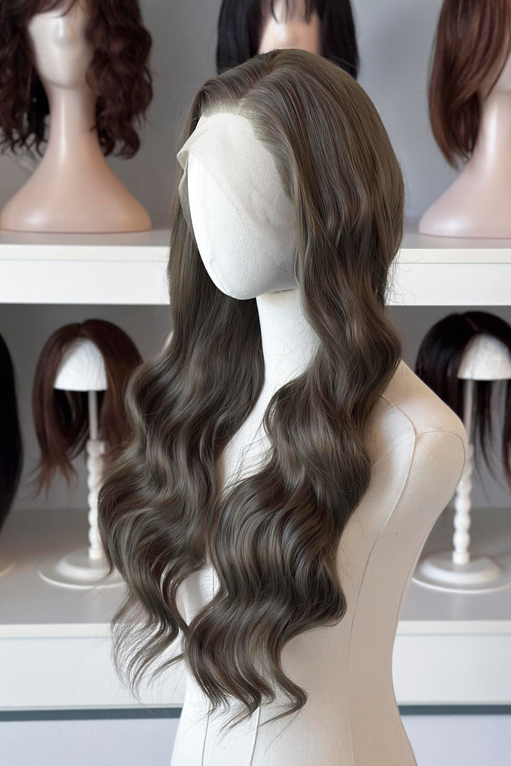 long wavy dark ash golden brown with green undertones wig in front of other mannequin heads with wigs on a shelf.