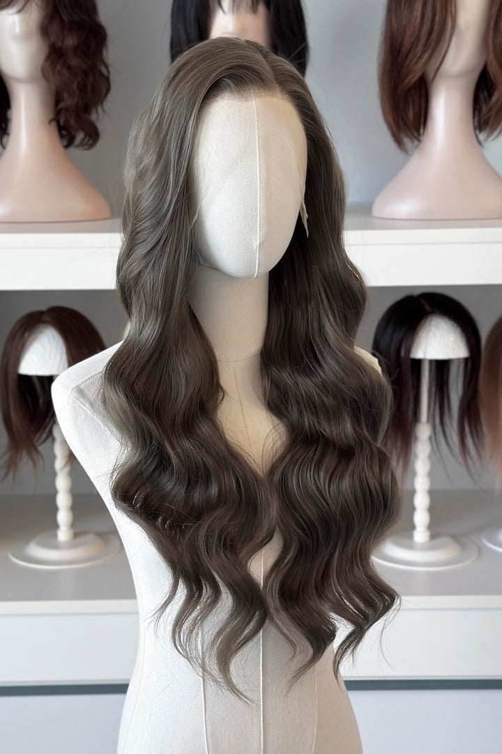 long wavy dark ash golden brown with green undertones wig in front of other mannequin heads with wigs on a shelf.
