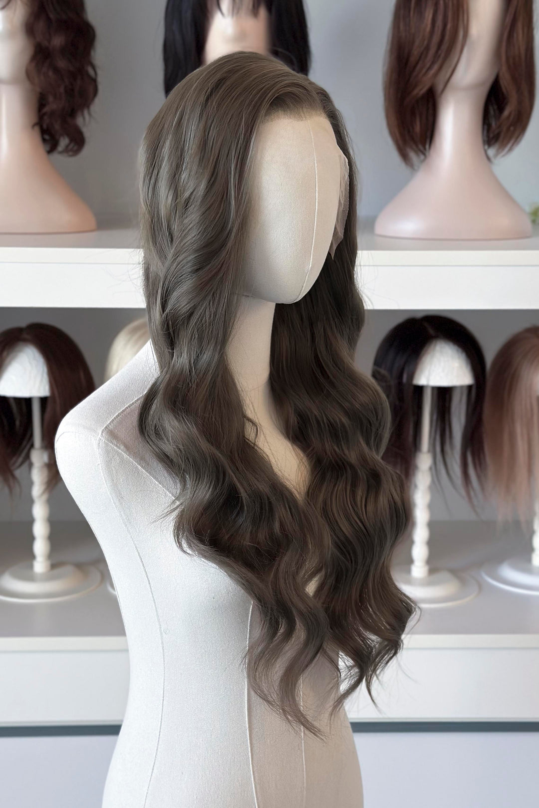 long wavy dark ash golden brown with green undertones wig in front of other mannequin heads with wigs on a shelf.