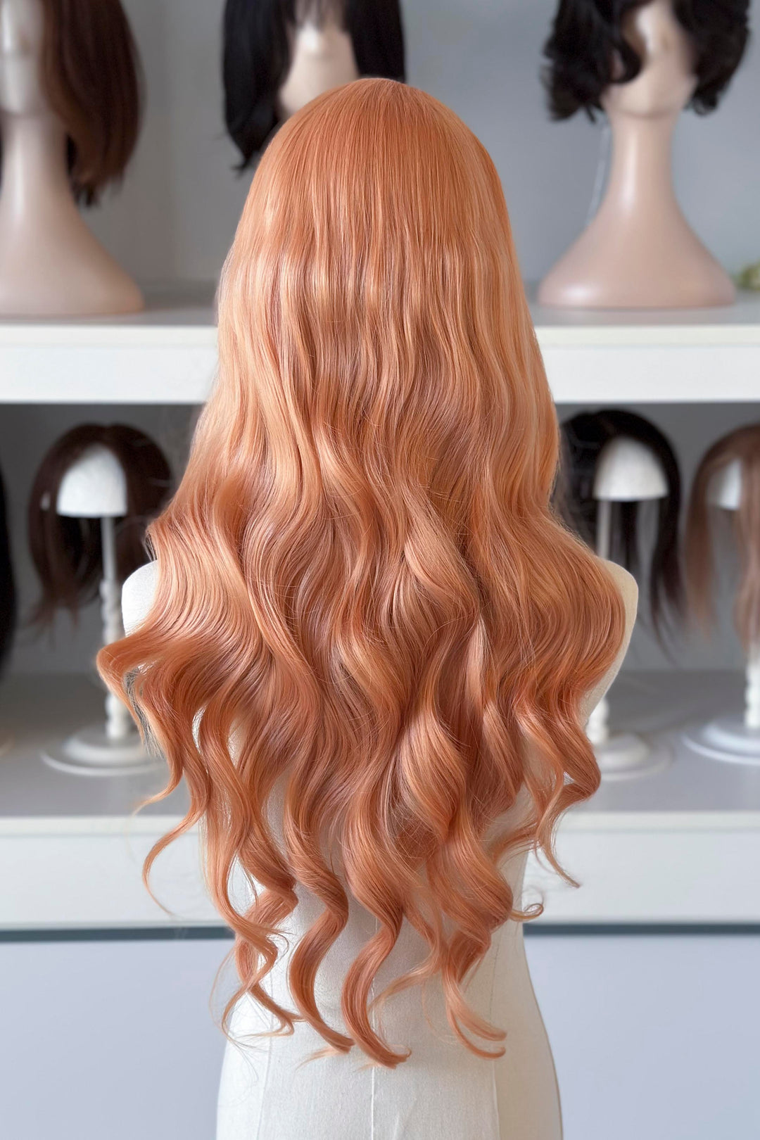 Long, wavy golden peach blonde wig on a mannequin head with blurred wig display in the background.