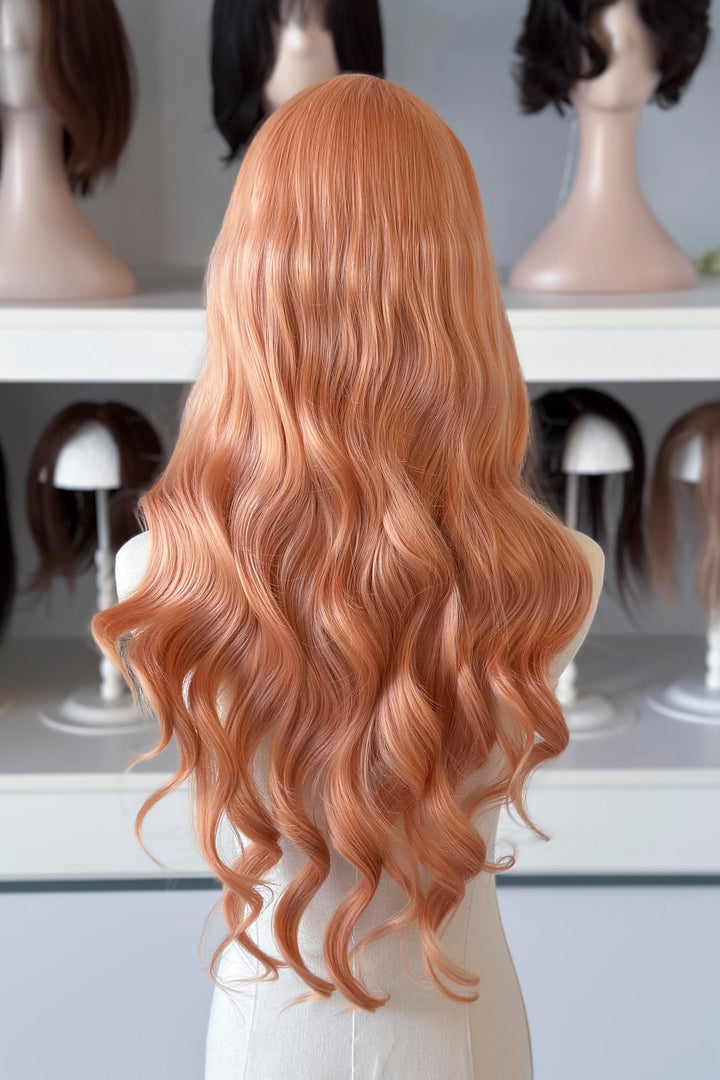 Long, wavy golden peach blonde wig on a mannequin head with blurred wig display in the background.