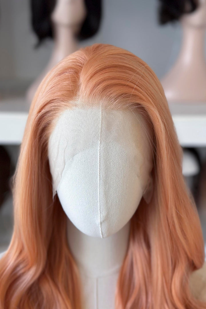 Long, wavy golden peach blonde wig on a mannequin head with blurred wig display in the background.