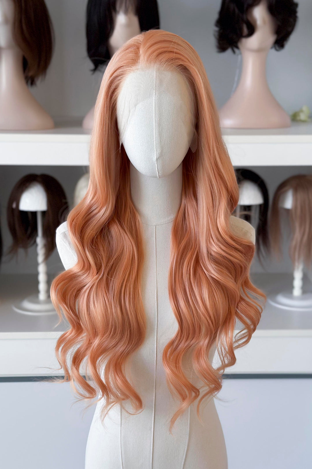 Long, wavy golden peach blonde wig on a mannequin head with blurred wig display in the background.