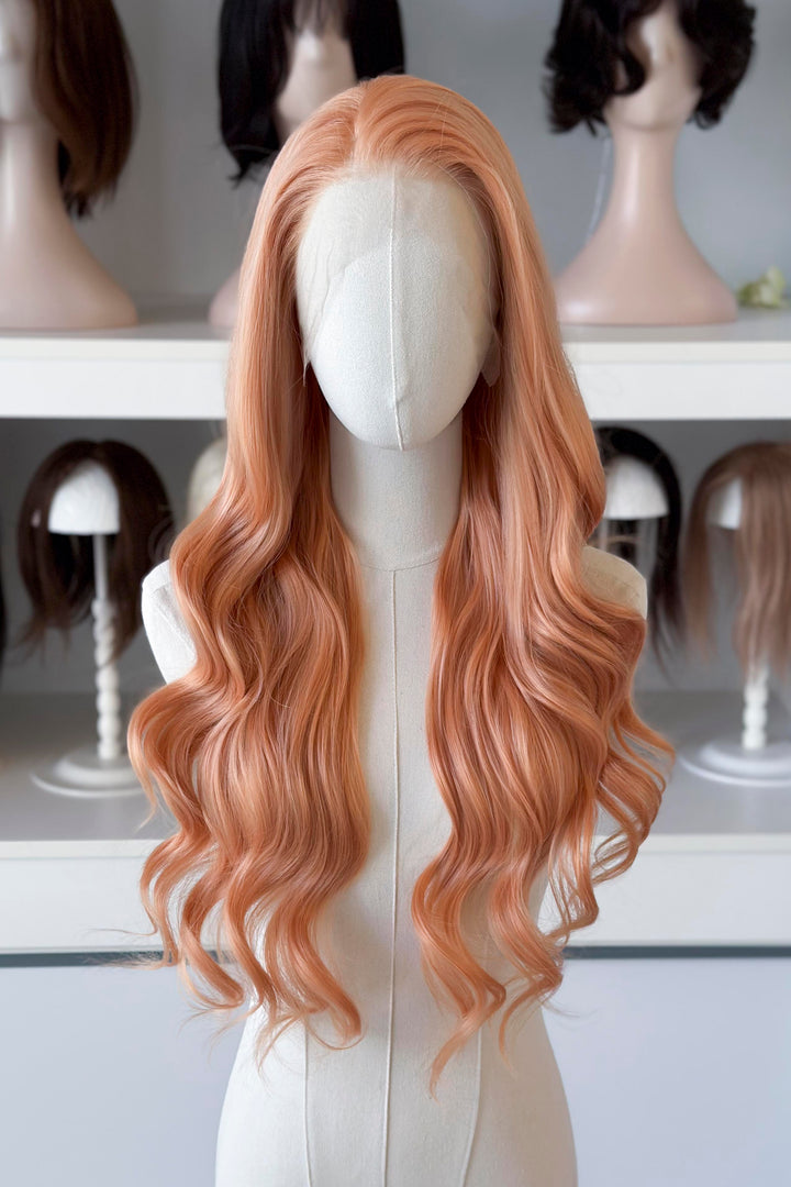 Long, wavy golden peach blonde wig on a mannequin head with blurred wig display in the background.