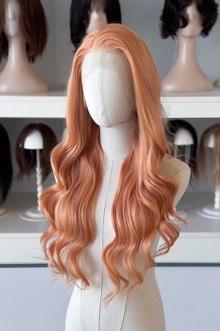 Long, wavy golden peach blonde wig on a mannequin head with blurred wig display in the background.