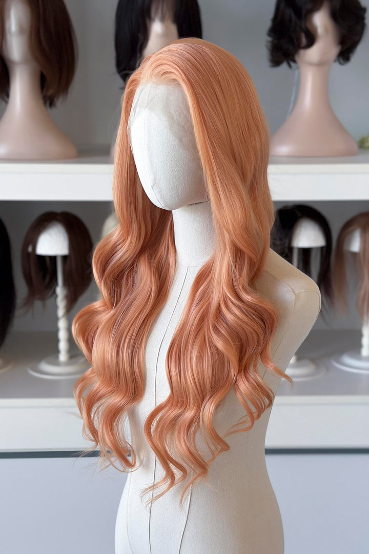 Long, wavy golden peach blonde wig on a mannequin head with blurred wig display in the background.