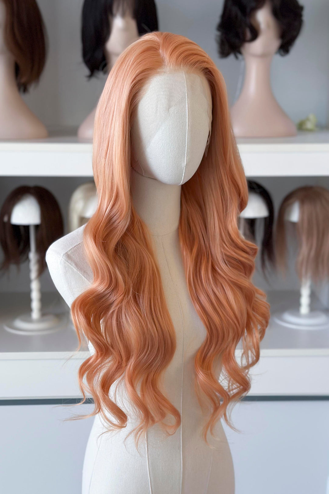 Long, wavy golden peach blonde wig on a mannequin head with blurred wig display in the background.