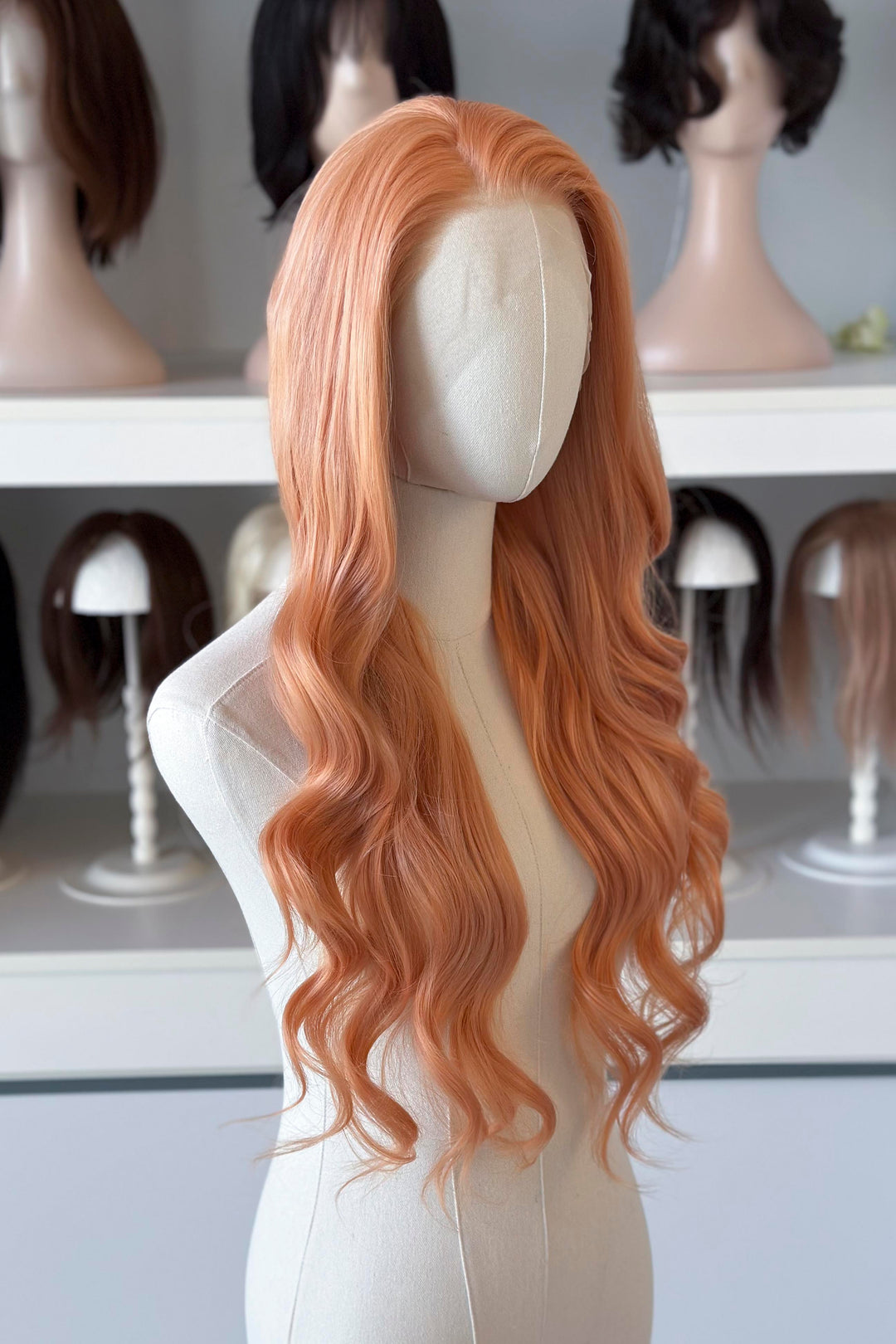 Long, wavy golden peach blonde wig on a mannequin head with blurred wig display in the background.