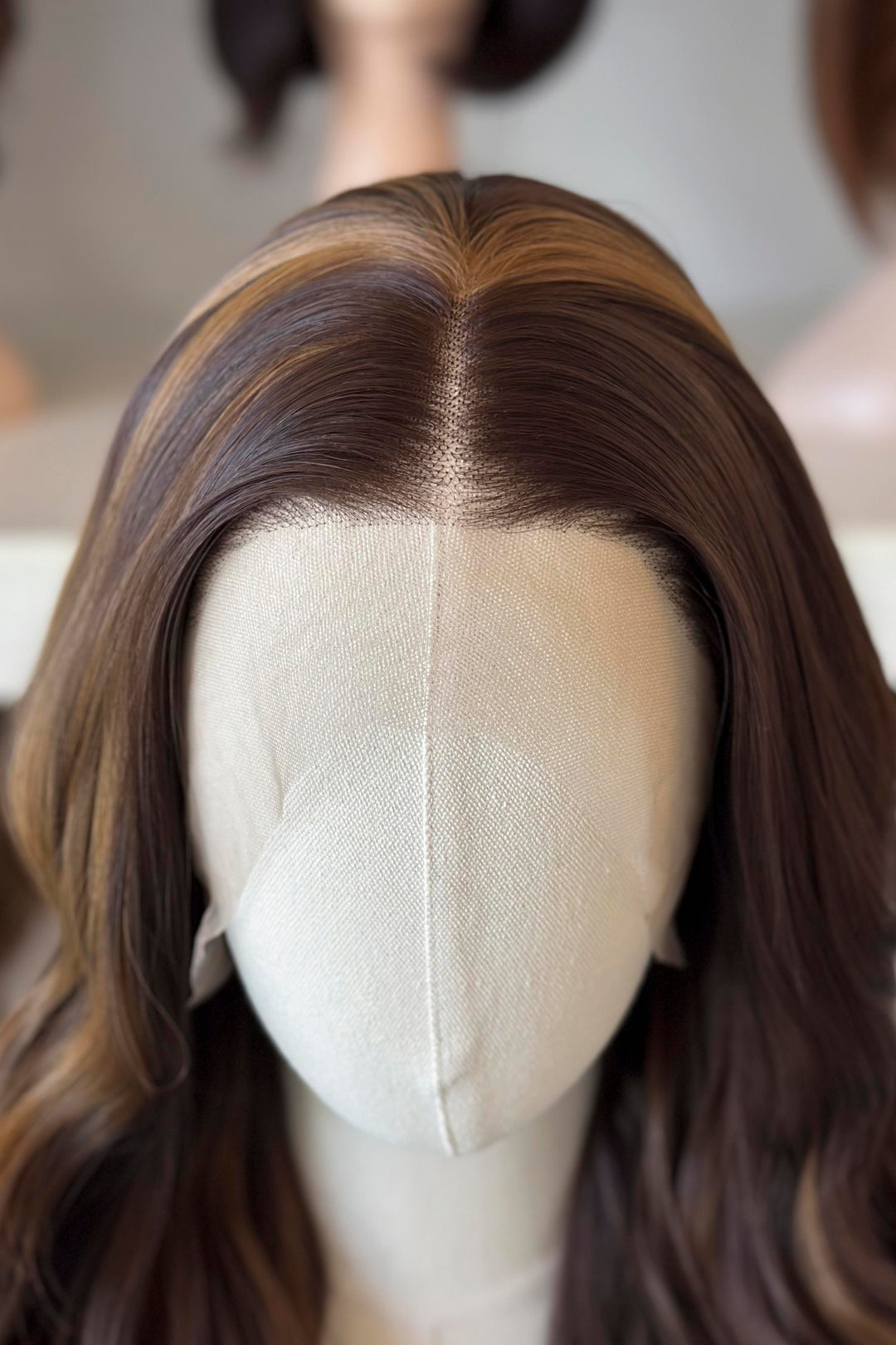 Brown wig with blonde highlights on a mannequin head with blurred wigs in the background