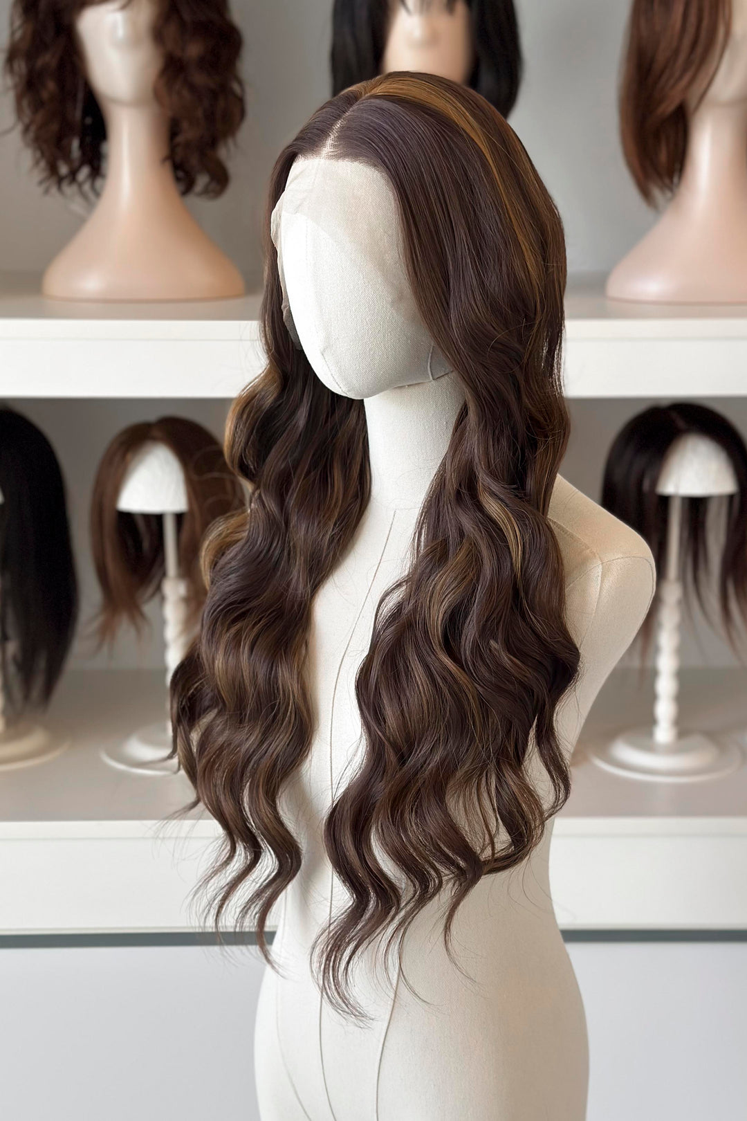 Brown wig with blonde highlights on a mannequin head with blurred wigs in the background