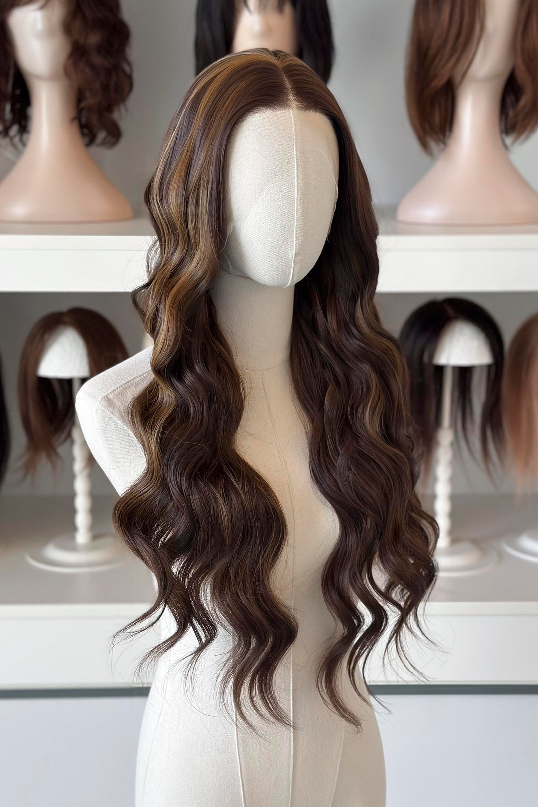 Brown wig with blonde highlights on a mannequin head with blurred wigs in the background