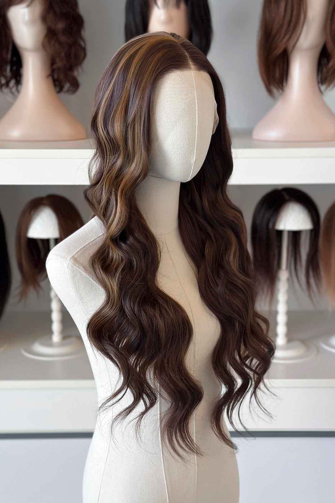 Brown wig with blonde highlights on a mannequin head with blurred wigs in the background