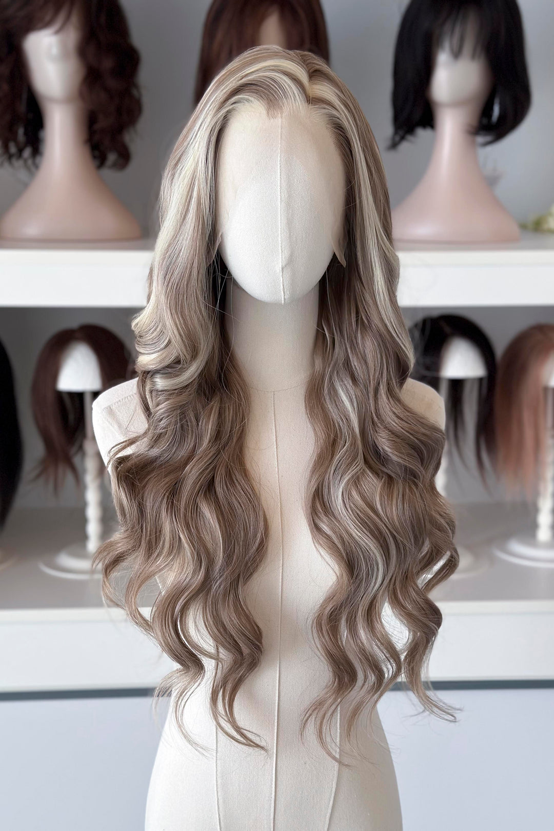 Ash brown hair with chunky blonde highlights wig on a mannequin head with blurred wigs in the background