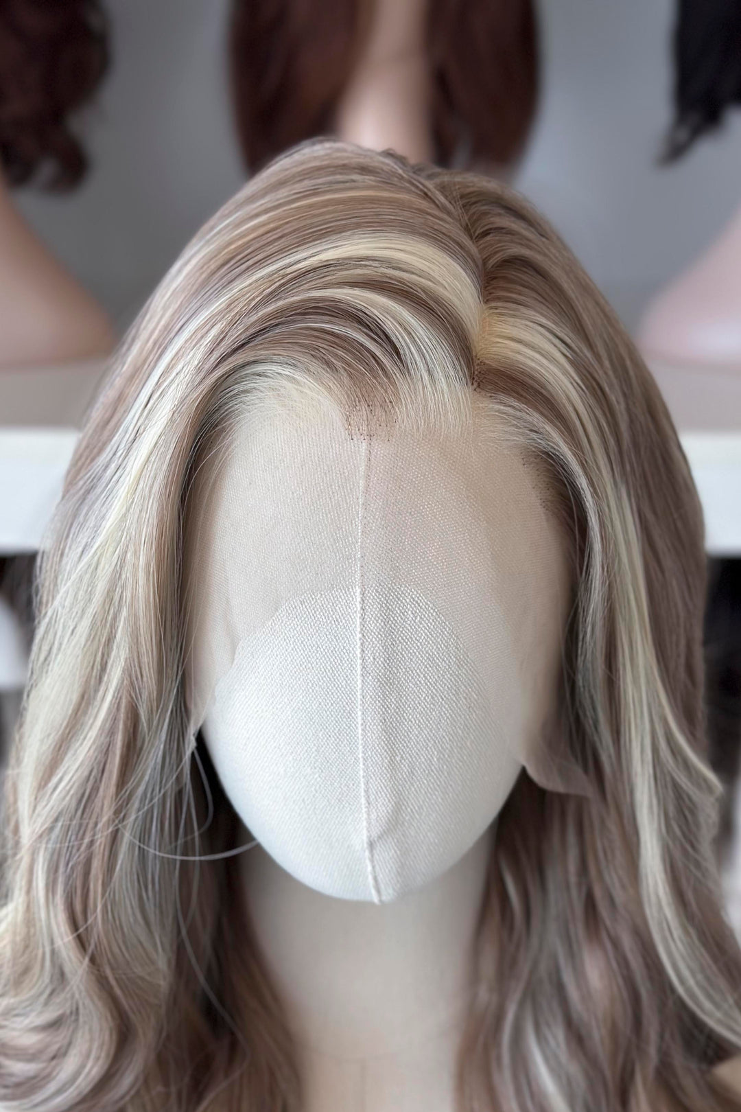 Ash brown hair with chunky blonde highlights wig on a mannequin head with blurred wigs in the background