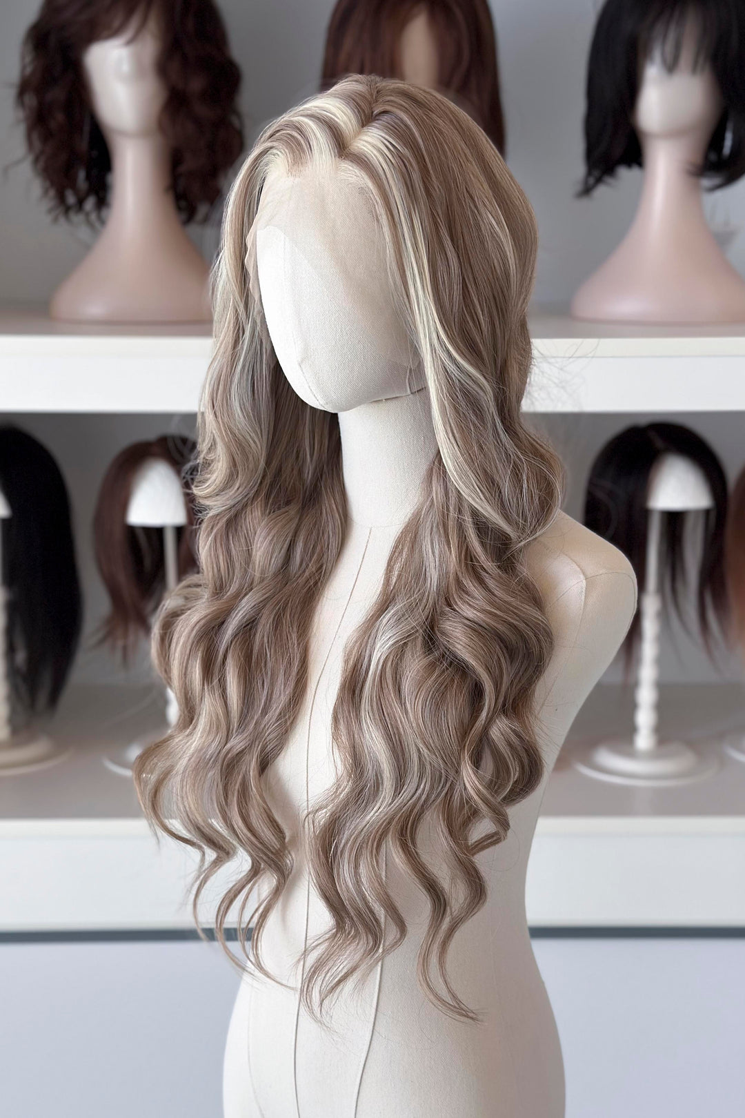 Ash brown hair with chunky blonde highlights wig on a mannequin head with blurred wigs in the background