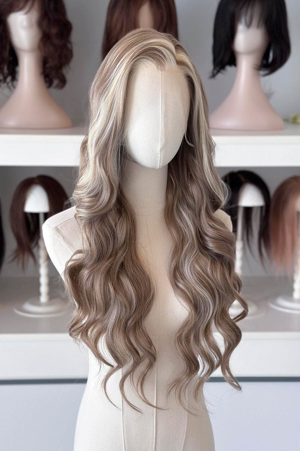 Ash brown hair with chunky blonde highlights wig on a mannequin head with blurred wigs in the background