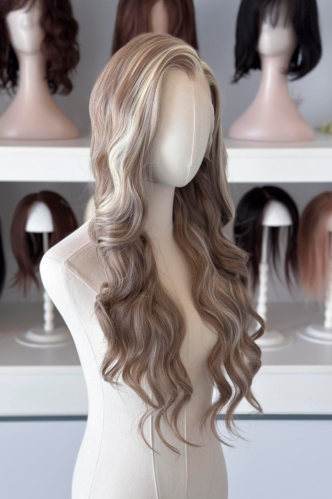 Ash brown hair with chunky blonde highlights wig on a mannequin head with blurred wigs in the background